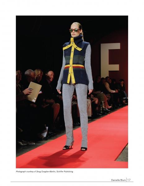 A model presents the latest look from "Emerging Fashion Designers 2" by Schiffer Publishing, Ltd., wearing a sleeveless navy vest with yellow accents and grey pants on a red runway as spectators watch, with a large letter E seen in the background.
