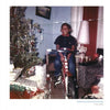 A smiling child sits on a red bike by wrapped presents and a Christmas tree in a cozy room. The vintage photo has a worn look, like the Original Gangster era. Product: Greg Batman Davis by Schiffer Publishing, Ltd.