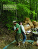 Two people stomp material outdoors on a tarp, surrounded by hay bales and supplies. In the upper left, a quote about cob stomping highlights the Schiffer Publishing, Ltd. product "Natural Building" and its natural building process.