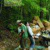 Two people stomp material outdoors on a tarp, surrounded by hay bales and supplies. In the upper left, a quote about cob stomping highlights the Schiffer Publishing, Ltd. product "Natural Building" and its natural building process.