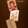 Three illustrated cards from "Today's Top Stationery Artists 2" by Schiffer Publishing, Ltd.: a girl making a heart, two girls bicycling, and a girl holding an umbrella over a fox on a brown background.