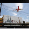 A commercial airplane flies low over a San Diego street above an upside-down mural, with power lines and a rail track in the foreground—a captivating urban scene featured in "Street Talking" by Schiffer Publishing, Ltd.