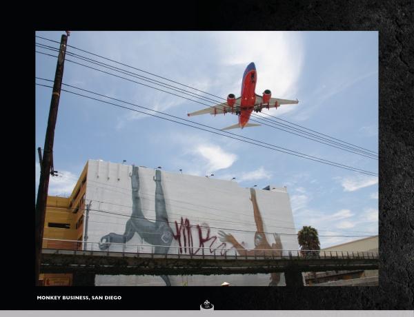 A commercial airplane flies low over a San Diego street above an upside-down mural, with power lines and a rail track in the foreground—a captivating urban scene featured in "Street Talking" by Schiffer Publishing, Ltd.