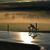 A cyclist rides a wet road at sunrise in Lancaster County, their reflection visible as mist drifts over fields—capturing the peaceful Amish life depicted in Faces of Lancaster County Postcards from Schiffer Publishing, Ltd.