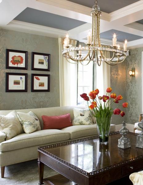 An inviting living room with a light sofa, red pillow, and vase of orange tulips on a polished table, inspired by "Showhouses 3" from Schiffer Publishing, Ltd., features wall art and a crystal chandelier overhead.