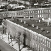 A black and white photo from "Bodine's City" by Schiffer Publishing, Ltd. depicts brick row houses, flat roofs, chimneys, and bare trees with a lone figure on the sidewalk, evoking the classic Baltimore style of Aubrey Bodine.