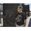 An elderly man in a veteran’s cap and black memorial T-shirt stands by a polished black granite war memorial, his commemorative tattoos inspired by War Paint from Schiffer Publishing, Ltd., honoring veterans alongside the inscribed names.