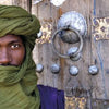 A man in a green turban and robe stands before an ornate wooden door of West African design; on the right, a panel features text about Timbuktu. From "Villages of West Africa" by Schiffer Publishing, Ltd.