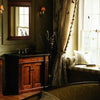 The Spa Bath by Schiffer Publishing, Ltd. showcases a warmly lit bathroom with a wooden vanity, mirror, wall sconce, vase with tall branches, and floral-curtained window seat—perfect for relaxing in spa-style comfort as sunlight streams in.