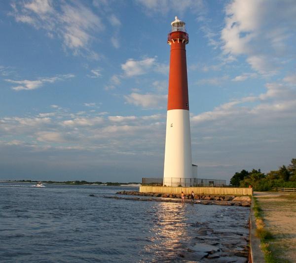 Barnegat Lighthouse Perspectives by Schiffer Publishing, Ltd. features the iconic red and white Barnegat Lighthouse on Long Beach Island, set beside calm water with sunlit reflections, rocky shoreline, fence, and a partly cloudy sky.