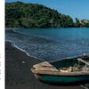 A man stands by a debris-filled wooden boat on a dark sandy beach, symbolizing contemporary Cuba, as green hills and buildings loom behind. An inset cross appears on the left. Product: Cuba's Evolution by Schiffer Publishing, Ltd.