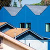 A row of bright blue modern homes with striped roofs from "Design with Love" by Schiffer Publishing, Ltd. stands out behind traditional tan and gray houses, illustrating innovation in community housing under a clear sky.