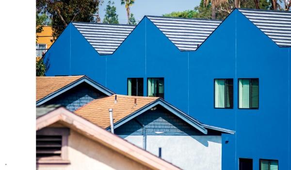 A row of bright blue modern homes with striped roofs from "Design with Love" by Schiffer Publishing, Ltd. stands out behind traditional tan and gray houses, illustrating innovation in community housing under a clear sky.