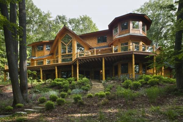 The Cedar Style by Schiffer Publishing, Ltd. features a spacious two-story log cabin with peaked roofs and a wraparound porch, nestled among trees on a landscaped hillside—highlighting timeless log home charm.