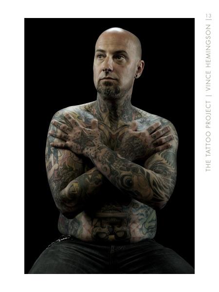 A bald, tattooed man with a goatee sits shirtless against a black background, showcasing intricate body art in this powerful image from "The Tattoo Project" by Schiffer Publishing, Ltd.