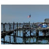 A pier with rustic wooden buildings, including a yellow one, stretches over calm water under a blue sky. Boats are docked nearby and an American flag flies—a scene from "Archipelago New York" by Schiffer Publishing, Ltd.