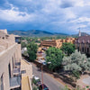 Experience Santa Fe’s vibrant culture with "Santa Fe Reflections" by Schiffer Publishing, Ltd.—featuring adobe buildings, a church steeple, tree-lined streets, and distant mountains under a partly cloudy sky.