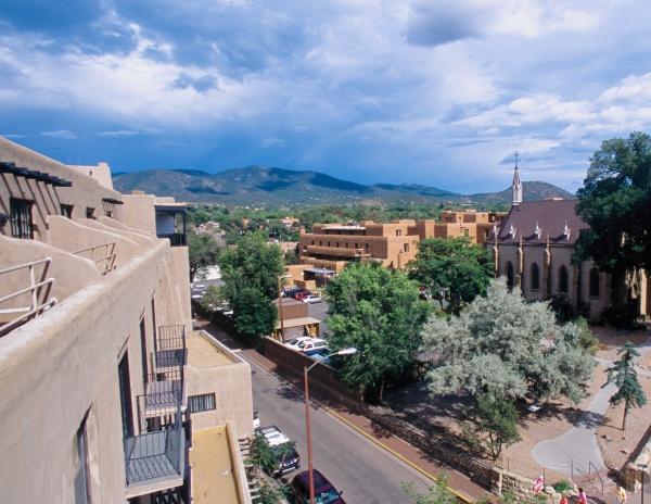 Experience Santa Fe’s vibrant culture with "Santa Fe Reflections" by Schiffer Publishing, Ltd.—featuring adobe buildings, a church steeple, tree-lined streets, and distant mountains under a partly cloudy sky.