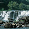 Capture the beauty of West Virginia with Schiffer Publishing, Ltd.: a wide waterfall flows over rocky ledges into a river, framed by lush trees and clear skies. Large rocks in the foreground create ideal spots for scenic photography.