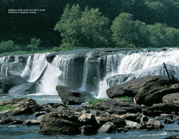 Capture the beauty of West Virginia with Schiffer Publishing, Ltd.: a wide waterfall flows over rocky ledges into a river, framed by lush trees and clear skies. Large rocks in the foreground create ideal spots for scenic photography.