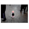 A small child in a pink hat and coat stands alone on a wet pavement among adults’ legs, evoking solitude—a quiet moment reminiscent of those in "Faces of Lancaster County" by Schiffer Publishing, Ltd.