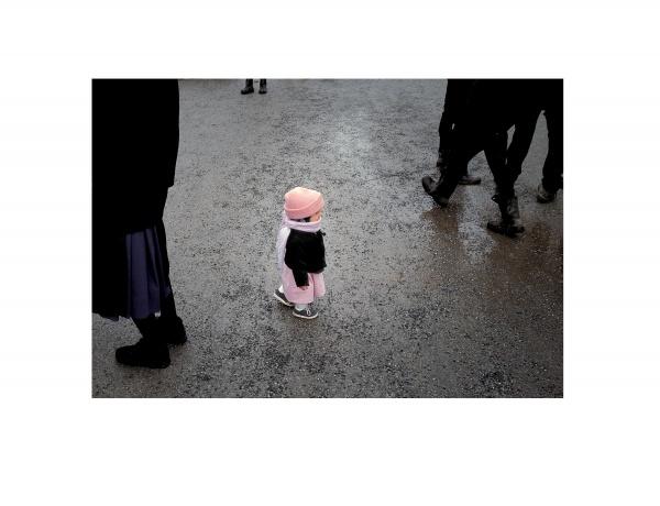 A small child in a pink hat and coat stands alone on a wet pavement among adults’ legs, evoking solitude—a quiet moment reminiscent of those in "Faces of Lancaster County" by Schiffer Publishing, Ltd.