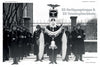 A black-and-white photo from "Black and Field Gray Uniforms of Himmler's SS, Vol. 1" by Schiffer Publishing, Ltd. shows Waffen-SS soldiers in formation in the snow with banners and standard; a building and iron fence are in the background.