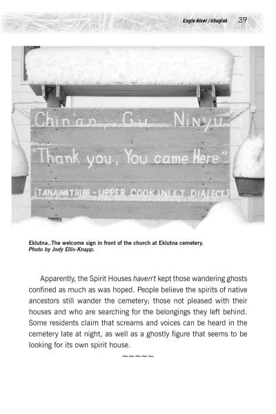 A wooden welcome sign dusted with snow hangs outside a white church at Eklutna cemetery, a location featured in "Ghosts of Alaska" by Schiffer Publishing, Ltd., where the sign displays both English and Native Upper Cook Inlet Dialect.