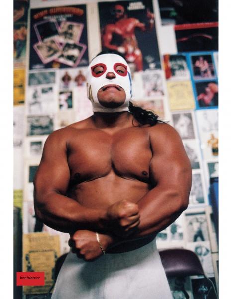 A muscular person in a white and red lucha libre mask flexes before a wall of Empire Wrestling Federation posters, embodying the spirit of pro wrestling celebrated in "Hard Boys and Bad Girls" by Schiffer Publishing, Ltd.