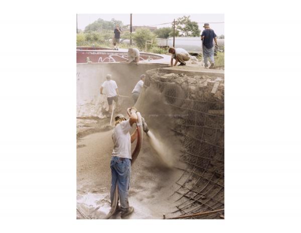 Workers spray concrete into a DIY skatepark bowl as others smooth the surface, capturing scenes featured in "FDR Skatepark: A Visual History" by Schiffer Publishing, Ltd., beneath an overcast sky.