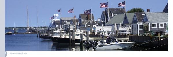 Boats dock at a harbor with gray shingled houses and American flags, under a clear sky. "Nantucket Vistas" by Schiffer Publishing, Ltd. captures this classic coastal scene in stunning photography.