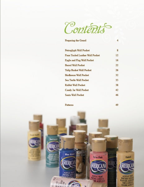 Several bottles of Americana acrylic paint in assorted colors sit on a light surface, with Schiffer Publishing, Ltd.'s "Creating Wall Pockets" book’s table of contents listing gourd painting and wall pocket projects visible in the background.