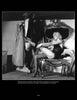 A woman in vintage lingerie adjusts her stockings before a mirror, surrounded by 1950s costumes—a scene echoing the glamorous burlesque of "Striptease Artists of the 1950s" from Schiffer Publishing, Ltd.