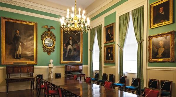 Inspired by American civic architecture, City Hall by Schiffer Publishing, Ltd. features a formal meeting room with a long wooden table, chandelier, green walls and curtains, empty chairs, portraits, and a bust.