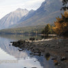 Experience the beauty of Glacier National Park with Schiffer Publishing, Ltd.—a rocky shore meets a calm lake that mirrors mountains and forests, with autumn’s orange and yellow hues evoking vintage postcard charm.