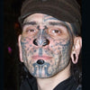 Featured in "Ink & Steel" by Schiffer Publishing, Ltd., this portrait captures a person with extensive facial tattoos, piercings (septum horn), chin implants, stretched earlobes, and a brown cap, facing the camera in dark clothing.