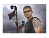 A man stands outdoors in sunglasses and a sleeveless shirt, revealing a large military tattoo on his upper arm, as three American flags wave behind him; featured on the cover of "War Paint" by Schiffer Publishing, Ltd.