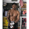 A serious male boxer in boxing shorts and gloves stands in a gym, his upper body sweaty. He wears a black headband, with fighter portraits and an American flag behind him—a scene featured in "Tough Guys" by Schiffer Publishing, Ltd.
