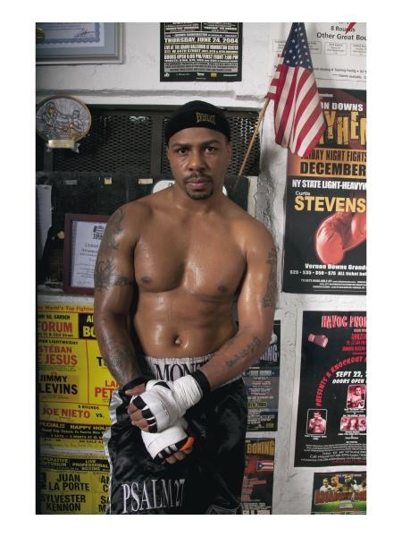 A serious male boxer in boxing shorts and gloves stands in a gym, his upper body sweaty. He wears a black headband, with fighter portraits and an American flag behind him—a scene featured in "Tough Guys" by Schiffer Publishing, Ltd.