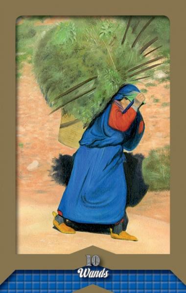 A person in blue and red struggles to carry a large bundle of sticks along a dirt path. The card, labeled 10 of Wands, is from the Tarot of the Moors by Schiffer Publishing, Ltd.