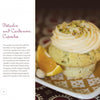 A pistachio and cardamom cupcake with swirled frosting and chopped pistachios sits on a plate beside a lemon wedge. The book "Dare to Bake!" by Schiffer Publishing, Ltd. is featured, highlighting creative cupcake recipes and flavors.