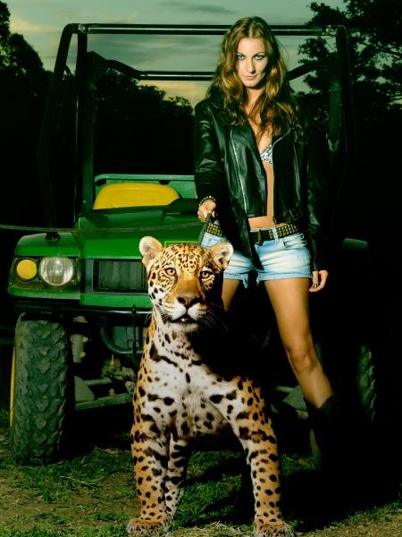 A woman in a black leather jacket and denim shorts stands outdoors with a jaguar on a leash and a green utility vehicle nearby at dusk, evoking the wild spirit of Paradise in Brazil by Schiffer Publishing, Ltd.