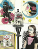 A woman with glasses stands in an art-filled room wearing a black cat shirt. She holds "Mommy's New Tattoo" by Schiffer Publishing, Ltd., surrounded by bold decor: panther tattoo art, a moon and stars portrait, skull sculpture, and lucky cat figure.