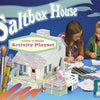 Two girls sit at a table coloring and assembling the Saltbox House by Schiffer Publishing. Art supplies and cut-outs are spread out, with "Saltbox House Activity Playset" text on a blue background featuring pencils and markers.