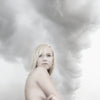 A blonde woman stands with her arms crossed, gazing ahead against swirling gray clouds—a moody, ethereal scene evocative of Iceland, featured in "Bare Landscapes" from Schiffer Publishing, Ltd.