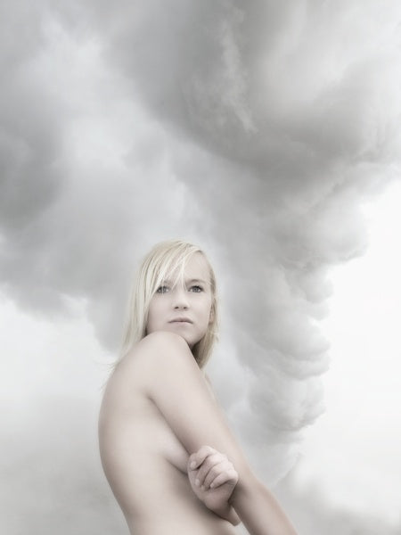 A blonde woman stands with her arms crossed, gazing ahead against swirling gray clouds—a moody, ethereal scene evocative of Iceland, featured in "Bare Landscapes" from Schiffer Publishing, Ltd.