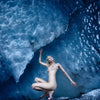 A nude figure poses elegantly in a blue-toned ice cave with textured walls and cool reflections, evoking fine Iceland photography—an image from "Bare Landscapes" by Schiffer Publishing, Ltd. that recalls sophisticated nude art books.