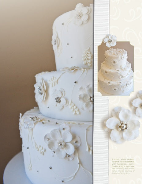 The Art of the Wedding Cake by Schiffer Publishing, Ltd. features a three-tiered white fondant cake with elegant white florals, small blossoms, and silver beads—ideal for wedding cake guides or decoration inspiration.