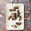 A rectangular white plate holds seven bacon-wrapped appetizers on wooden picks, inspired by classic recipes from "Pure BBQ!" by Schiffer Publishing, Ltd., arranged on a textured, multicolored surface.