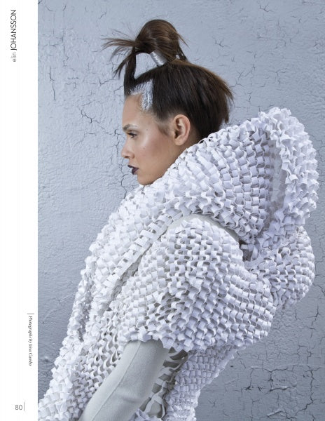 Against a gray, cracked wall, a person with styled brown hair and silver accents models an elaborate white outfit with a sculptural collar—showcasing innovative designs from "Emerging Fashion Designers 3" by Schiffer Publishing, Ltd.